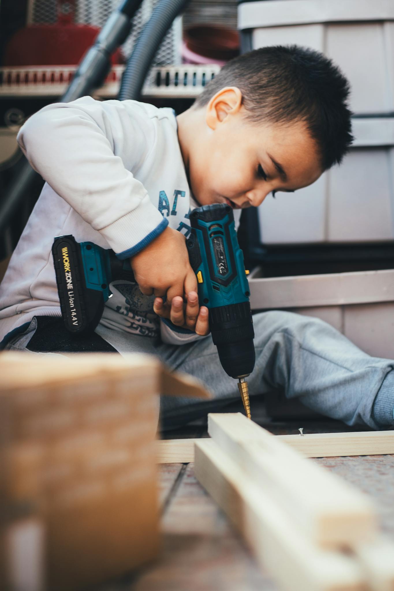 A young boy learning carpentry indoors, using a drill on wood.